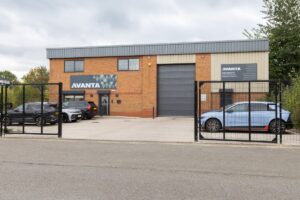 Industrial brick building with Avanta signage, a large gray roller shutter, and a gated parking lot with several cars visible.