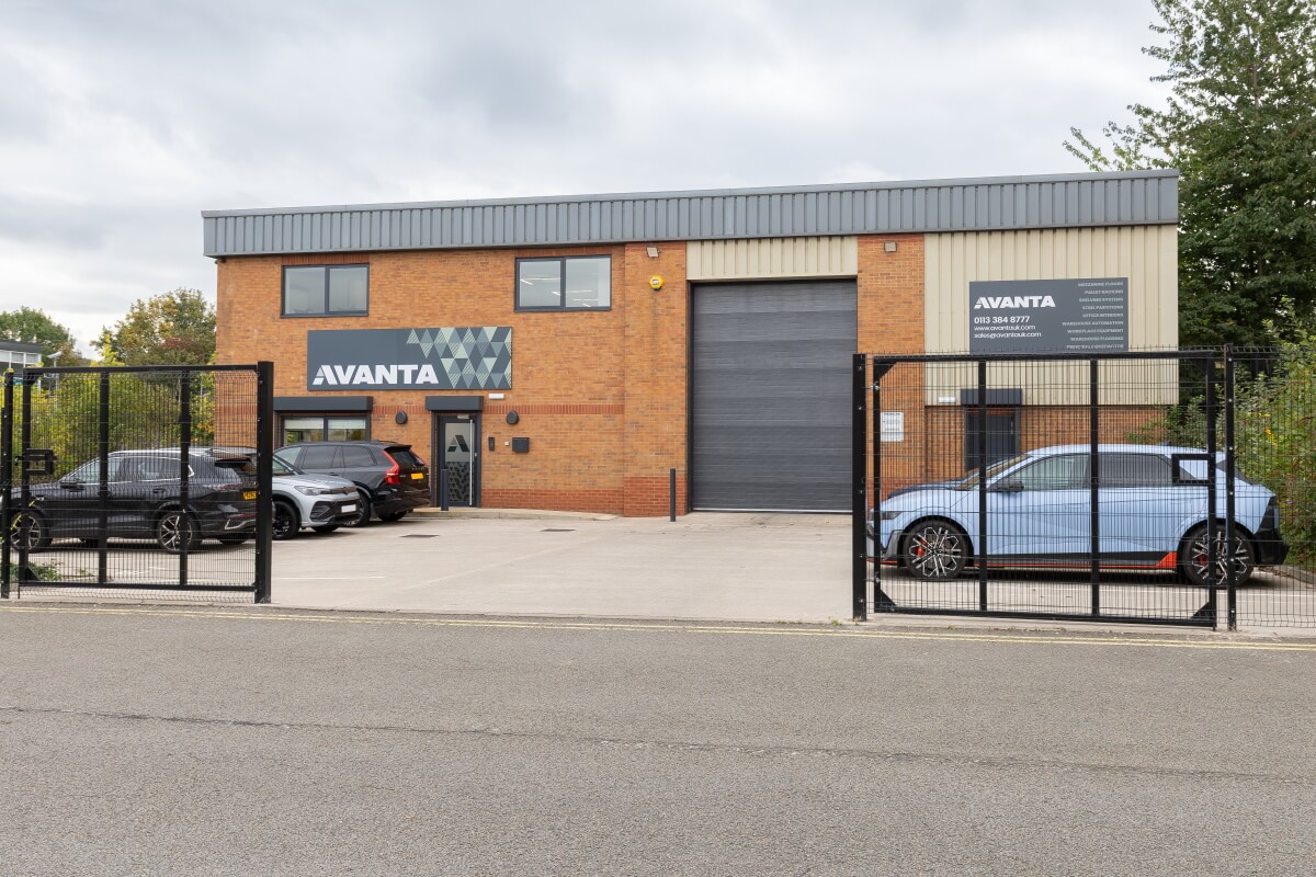 Industrial brick building with Avanta signage, a large gray roller shutter, and a gated parking lot with several cars visible.