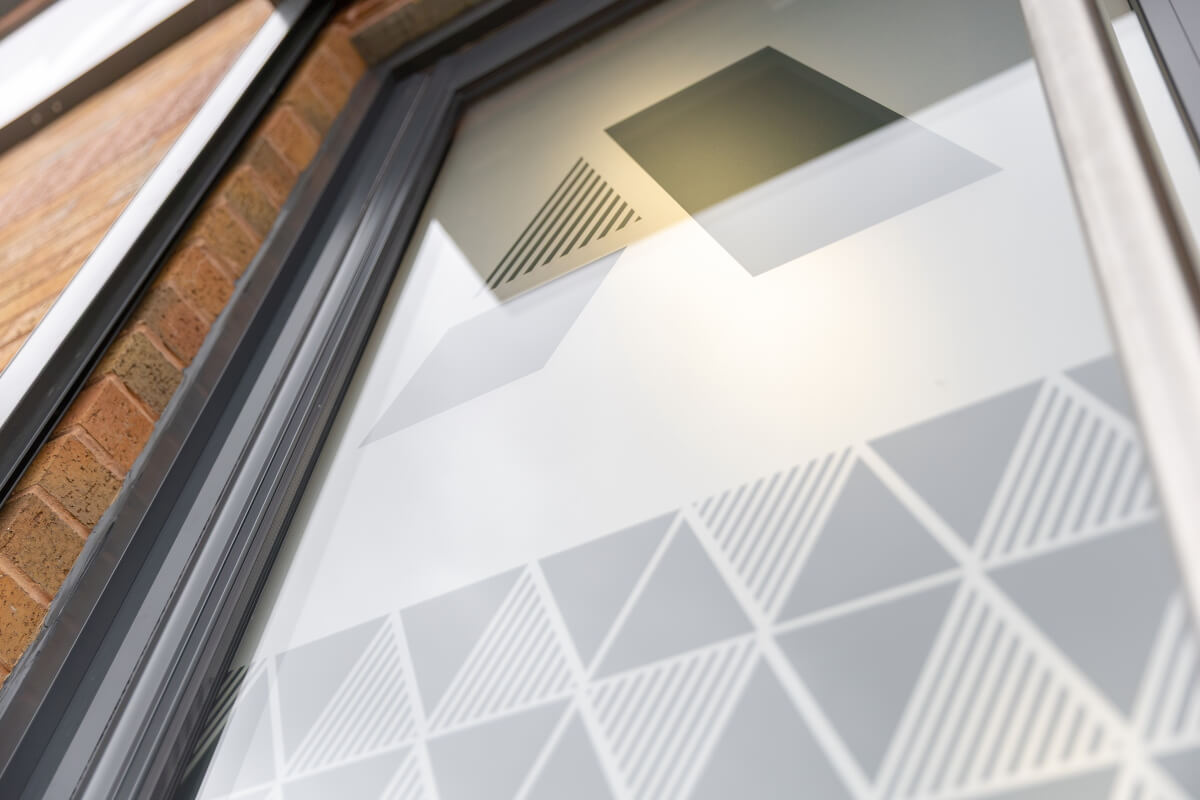 Storefront window with frosted geometric pattern of triangles and stripes on glass, brick wall visible in frame.