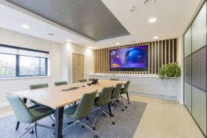 Modern conference room with a long wooden table, eight green swivel chairs, and a wall-mounted display framed by wooden slats on a decorative wall.