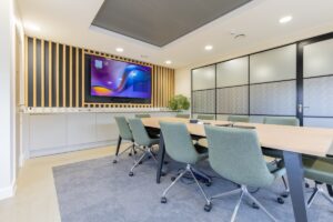 Modern conference room with a long wooden table, green swivel chairs, and a wall-mounted TV framed by vertical wooden slats.