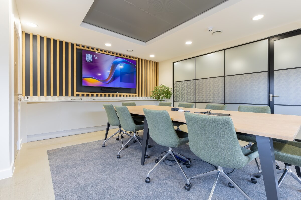 Modern conference room with a long wooden table, green swivel chairs, and a wall-mounted TV framed by vertical wooden slats.