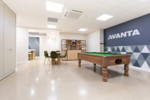 Open-plan office lounge with a wooden pool table, green felt, and a blue Avanta logo wall nearby.