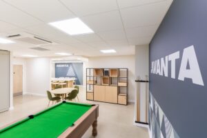 Bright coworking space with a green-felt pool table in the foreground, round table and chairs, and a blue wall featuring the AVANTA logo behind storage shelves.