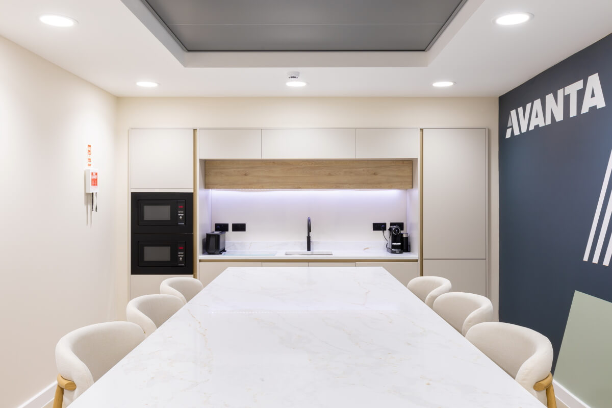 Long white marble conference table with cream chairs in a bright office kitchenette, featuring ovens, sink, and coffee station against an Avanta-branded wall.