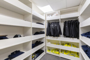 White walk-in storage closet with coats hanging on a rail and shelves holding high-visibility yellow vests.