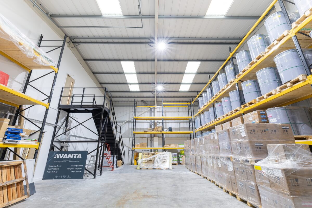 Wide view of a warehouse interior with tall yellow racks, palletized goods on both sides, and a metal staircase in the center area with wrapped pallets on the floor.