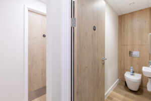 Public restroom with wooden doors; one door open revealing a toilet stall in a bright, modern washroom