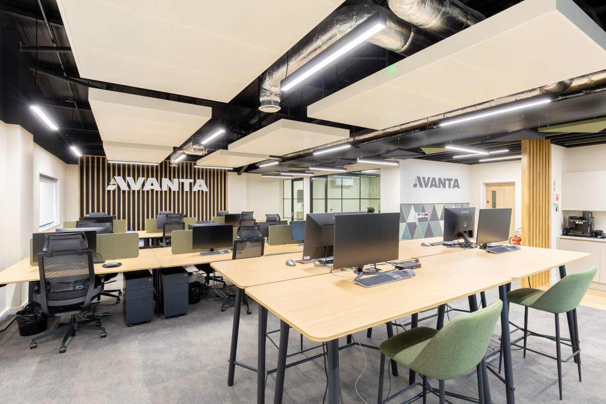 Open-plan office with shared wooden tables, computer workstations, and AVANTA logo on a striped wooden wall above the desks