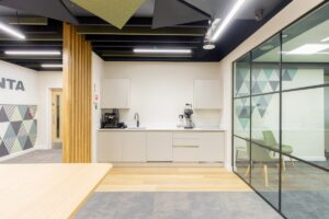 Modern office kitchenette with coffee machine, kettle, sink and light gray cabinets along a white counter. Nearby glass-walled meeting room with geometric wall pattern.