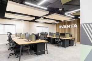 Open-plan modern office workspace with long desks, ergonomic chairs, partition screens, and a wood slat wall with AVANTA branding on the back wall.