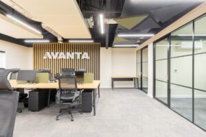 Open-plan office with desks, mesh chairs, and green acoustic panels, featuring an Avanta sign on a wooden slat wall toward the back.