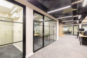 Open-plan office with glass-walled meeting rooms, gray carpeting, and geometric ceiling panels.